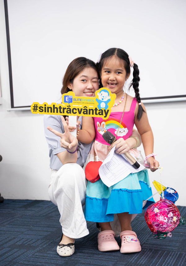 Bé gái cười tươi bên cô giáo cùng bảng hashtag sinh trắc vân tay và thành phẩm lồng đèn.