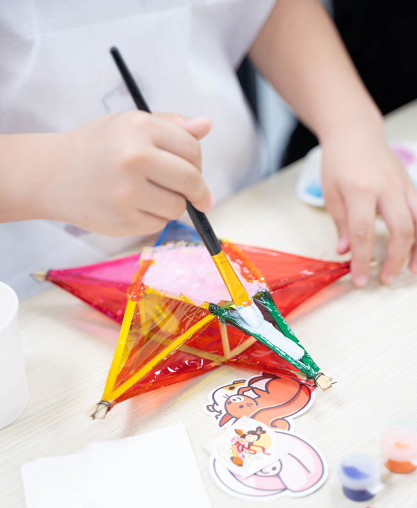 Cận cảnh bàn tay bé dùng cọ tô màu trang trí lồng đèn ngôi sao nhiều màu sắc.