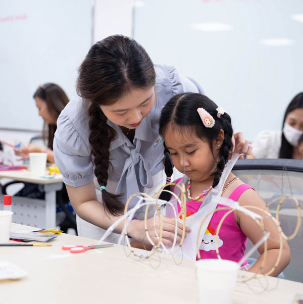 Hướng dẫn viên tỉ mỉ chỉ dẫn bé gái dán giấy màu lên khung lồng đèn thủ công.